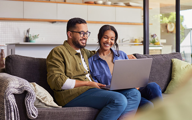 a couple sitting on a couch at home smiling and looking at their laptop